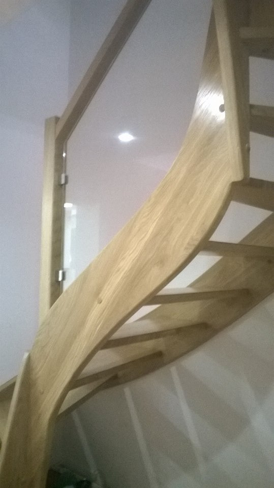 Klatka schodowa z jasnego drewna z zakrzywioną balustradą i szklanym panelem ochronnym, widok z dołu.