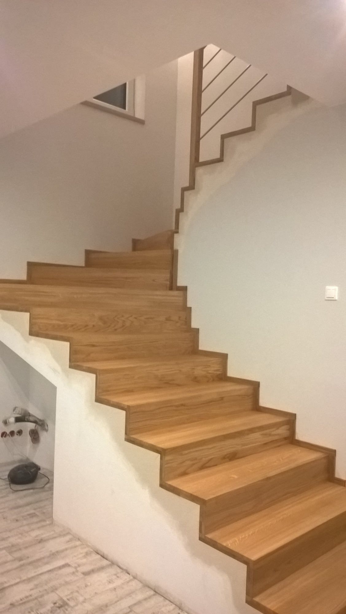 Drewniane schody z minimalistyczną, metalową balustradą w nowoczesnym domu. Widoczny fragment trójkątnego okna w ścianie. Pod schodami znajduje się schowek z odkurzaczem i butami.