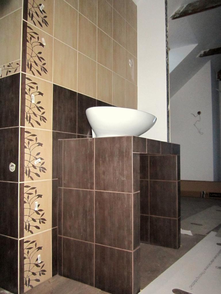 Łazienka w trakcie wykańczania: umywalka nablatowa na postumencie z brązowych płytek, ściany wyłożone płytkami w odcieniach beżu i brązu z motywem roślinnym.