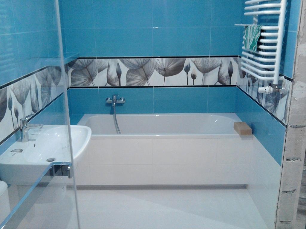 Wykończona łazienka z błękitnymi płytkami, dekoracyjnym pasem z motywem roślinnym, białą wanną obudowaną płytkami, umywalką i grzejnikiem drabinkowym z ręcznikiem.