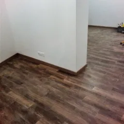 Wnętrze po remoncie z nową, ciemną podłogą imitującą drewno, białe ściany z gniazdkiem elektrycznym i widocznym fragmentem drugiego pomieszczenia z narzędziami w tle.