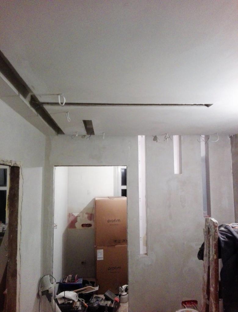 Wnętrze w trakcie prac: surowe ściany po nałożeniu gładzi, widoczne profile pod zabudowę sufitu, kartony z materiałami budowlanymi oraz narzędzia w nieładzie.