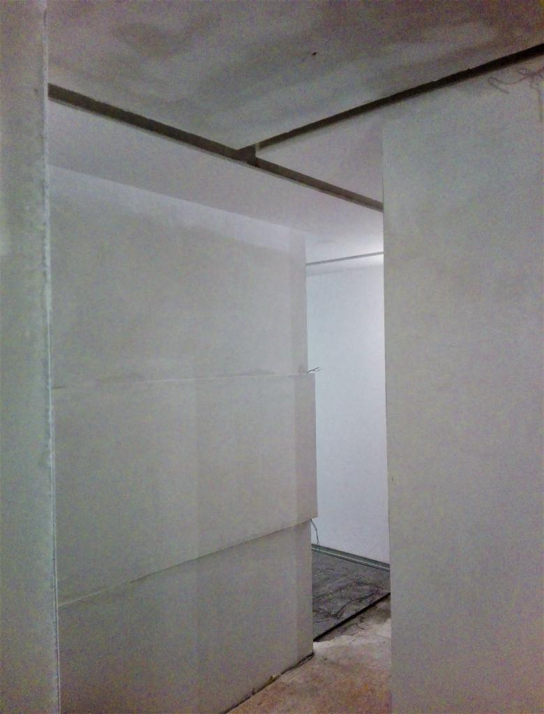 Wnętrze w trakcie prac: surowy stan ścian po szpachlowaniu, widoczne łączenia płyt kartonowo-gipsowych na suficie oraz fragment podłogi zabezpieczonej folią.