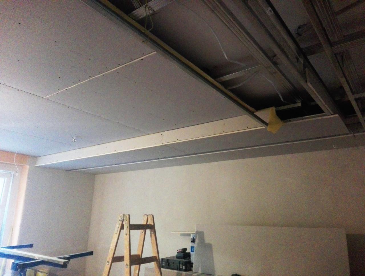 Montaż sufitu podwieszanego z płyt kartonowo-gipsowych, widoczne metalowe profile, fragment niezabudowanej przestrzeni i drabina malarska w pomieszczeniu.