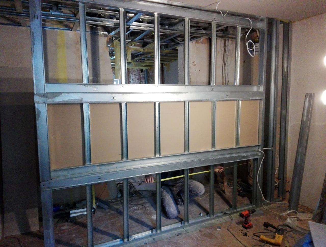 Konstrukcja metalowa z płytami kartonowo-gipsowymi w trakcie montażu, widoczny pracownik mierzący odległość taśmą mierniczą, narzędzia leżą na podłodze, surowy stan wnętrza.