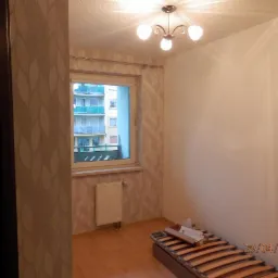 Pokój w trakcie przygotowań do remontu: ściany częściowo pomalowane na biało, tapeta w liście na jednej ścianie, drewniana podłoga, okno z widokiem na blok, częściowo złożony stelaż łóżka na podłodze.