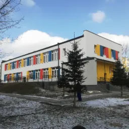 Dwupiętrowy budynek po renowacji elewacji, z białymi ścianami i kolorowymi panelami (żółty, czerwony, niebieski) wokół okien, widoczne drzewa iglaste i resztki śniegu na trawniku.