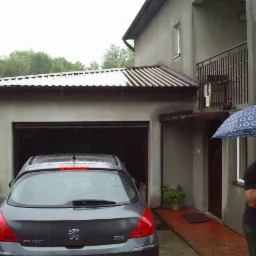 Szary dom jednorodzinny z garażem w deszczowy dzień, zaparkowany szary samochód Peugeot 308, mężczyzna z parasolem w tle.