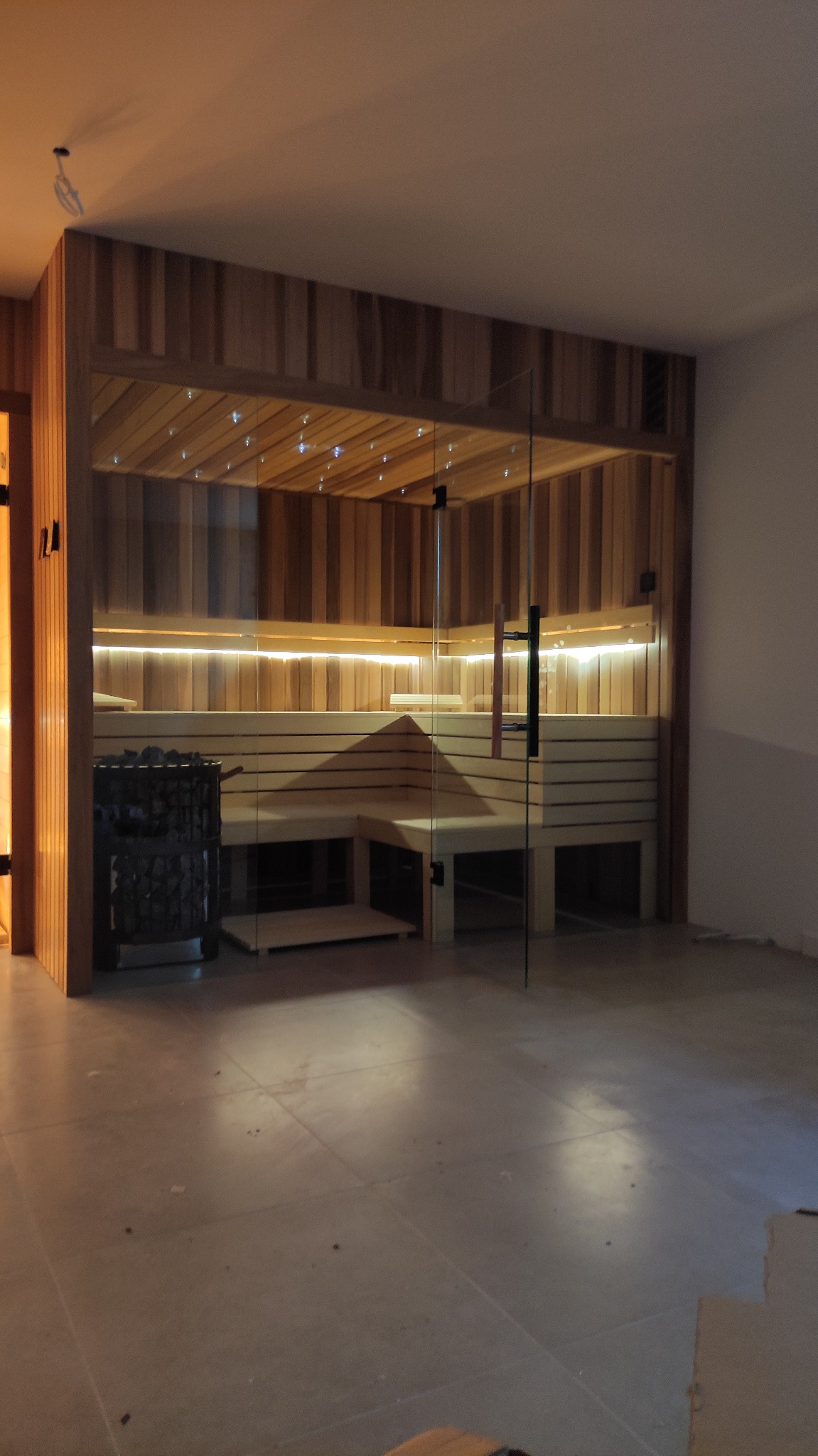 Nowoczesna kabina z drewna z przeszklonymi drzwiami, oświetleniem LED pod ławkami i piecem na kamienie w rogu.