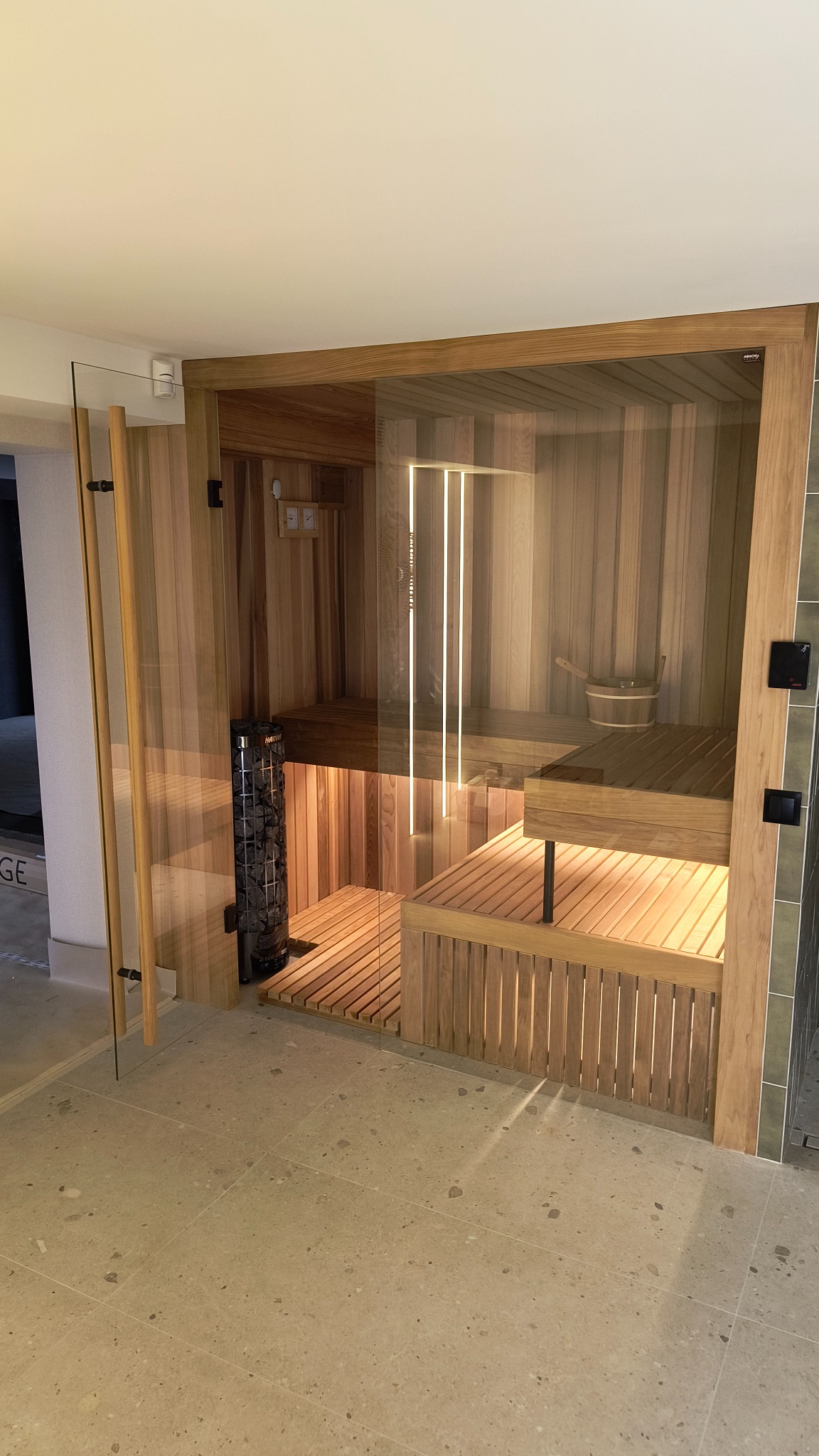 Nowoczesna, drewniana kabina z przeszklonymi drzwiami, wyposażona w piec z kamieniami i oświetlenie LED, z widocznym wiadrem i chochlą.