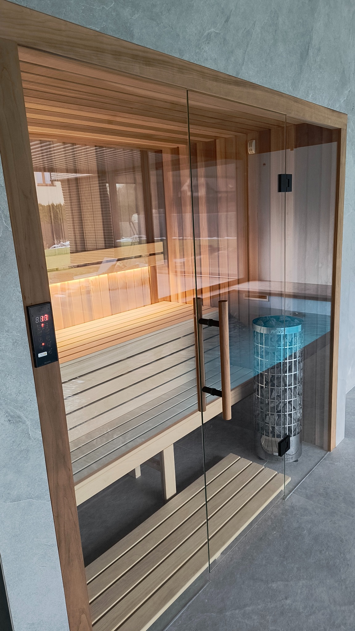 Szklane drzwi do sauny z drewnianą ramą, widoczne wnętrze z ławkami, piec z kamieniami i panel sterowania temperaturą.