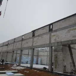 Wznoszenie ścian z betonu komórkowego na budowie hali, widoczny żuraw budowlany i materiały izolacyjne.