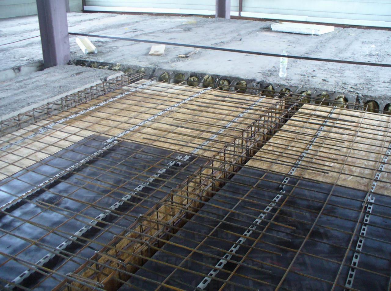 Zbrojenie stropu w trakcie prac budowlanych, widoczne siatki z prętów stalowych, płyty szalunkowe i izolacja termiczna w otworach, na tle surowego betonu i metalowej konstrukcji.