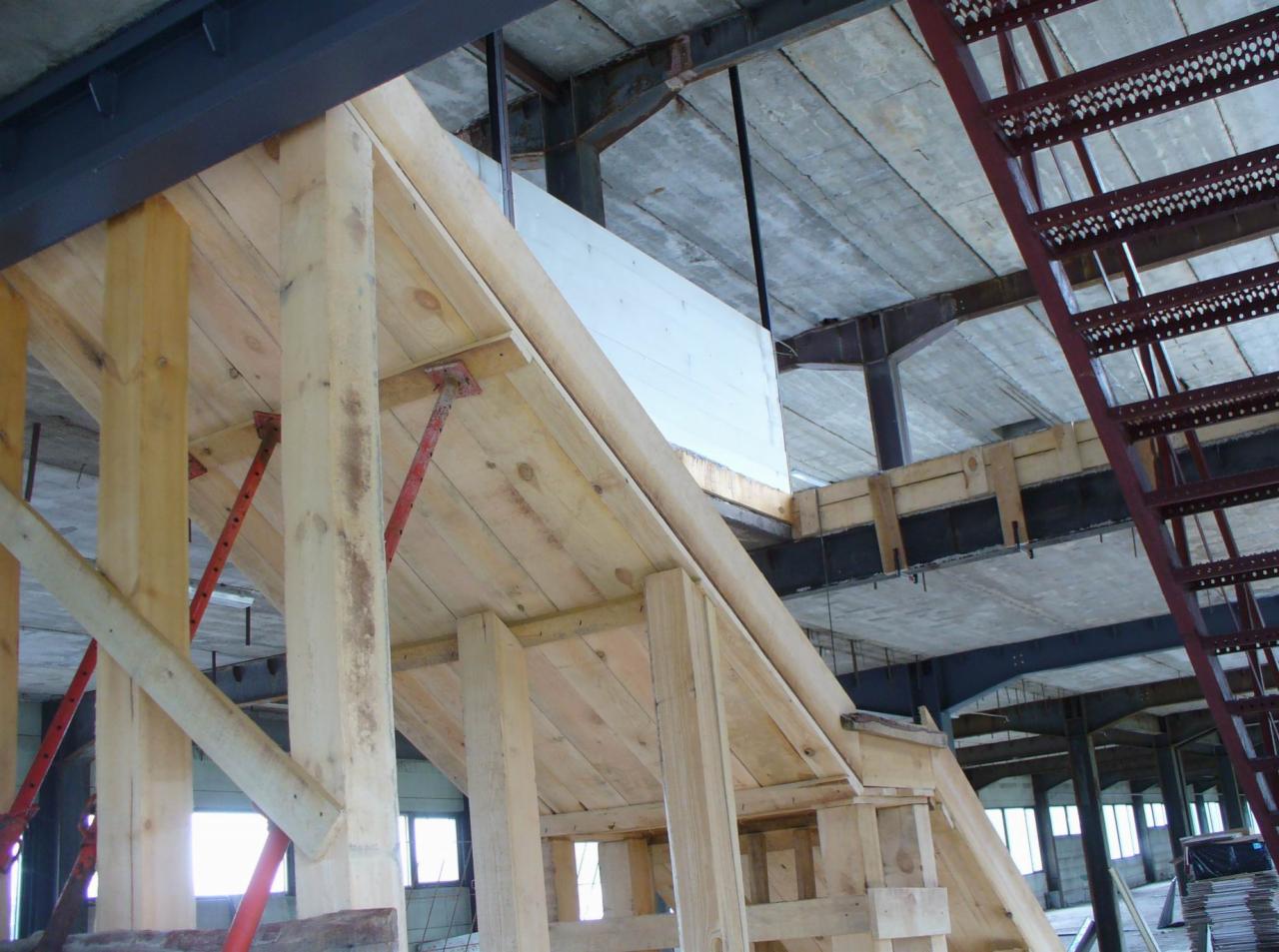 Szczegółowe ujęcie drewnianego szalunku pod schody w surowym stanie budynku, z widocznymi stalowymi belkami konstrukcyjnymi i metalowymi podporami.