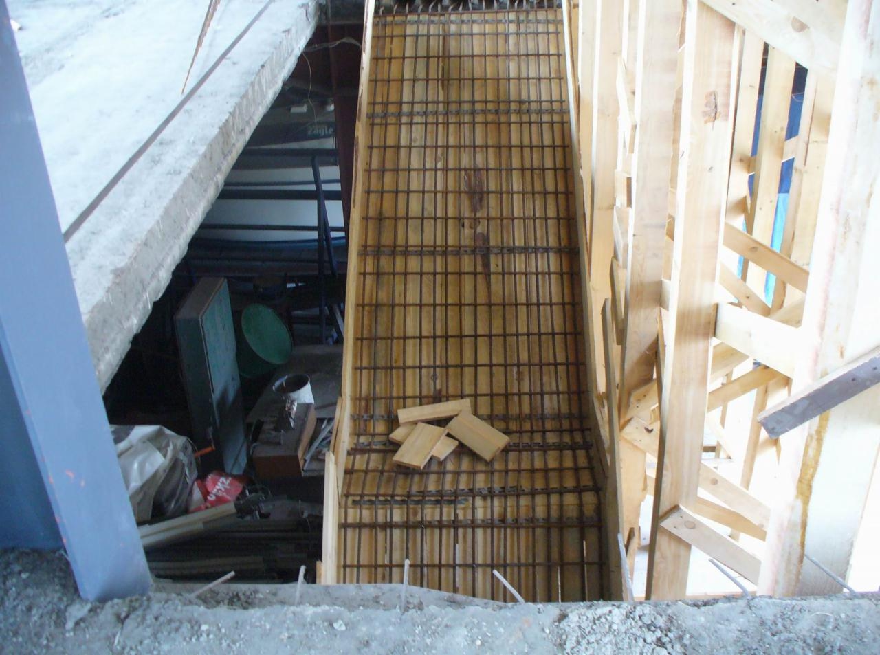 Zbrojenie schodów w drewnianym szalunku, widoczne elementy konstrukcji nośnej i fragment pomieszczenia z wyposażeniem budowlanym.