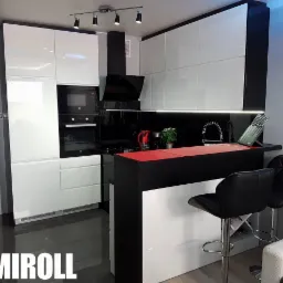 Nowoczesna kuchnia z białymi, lakierowanymi frontami szafek, czarnym blatem i barkiem z czerwonym akcentem, oświetlona punktowymi lampami i taśmą LED pod szafkami wiszącymi. Dwa czarne krzesła...