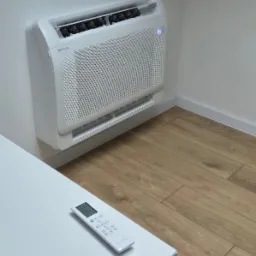 Biała jednostka klimatyzacji podłogowej zamontowana na ścianie w rogu pomieszczenia, obok leży pilot na białym blacie, widoczna drewniana podłoga.