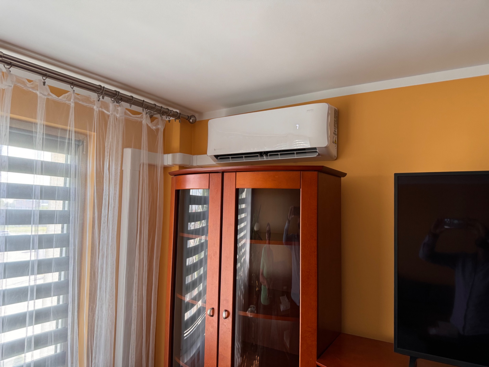 Biały klimatyzator ścienny zamontowany nad drewnianym kredensem w pokoju z pomarańczowymi ścianami i firankami w oknie.
