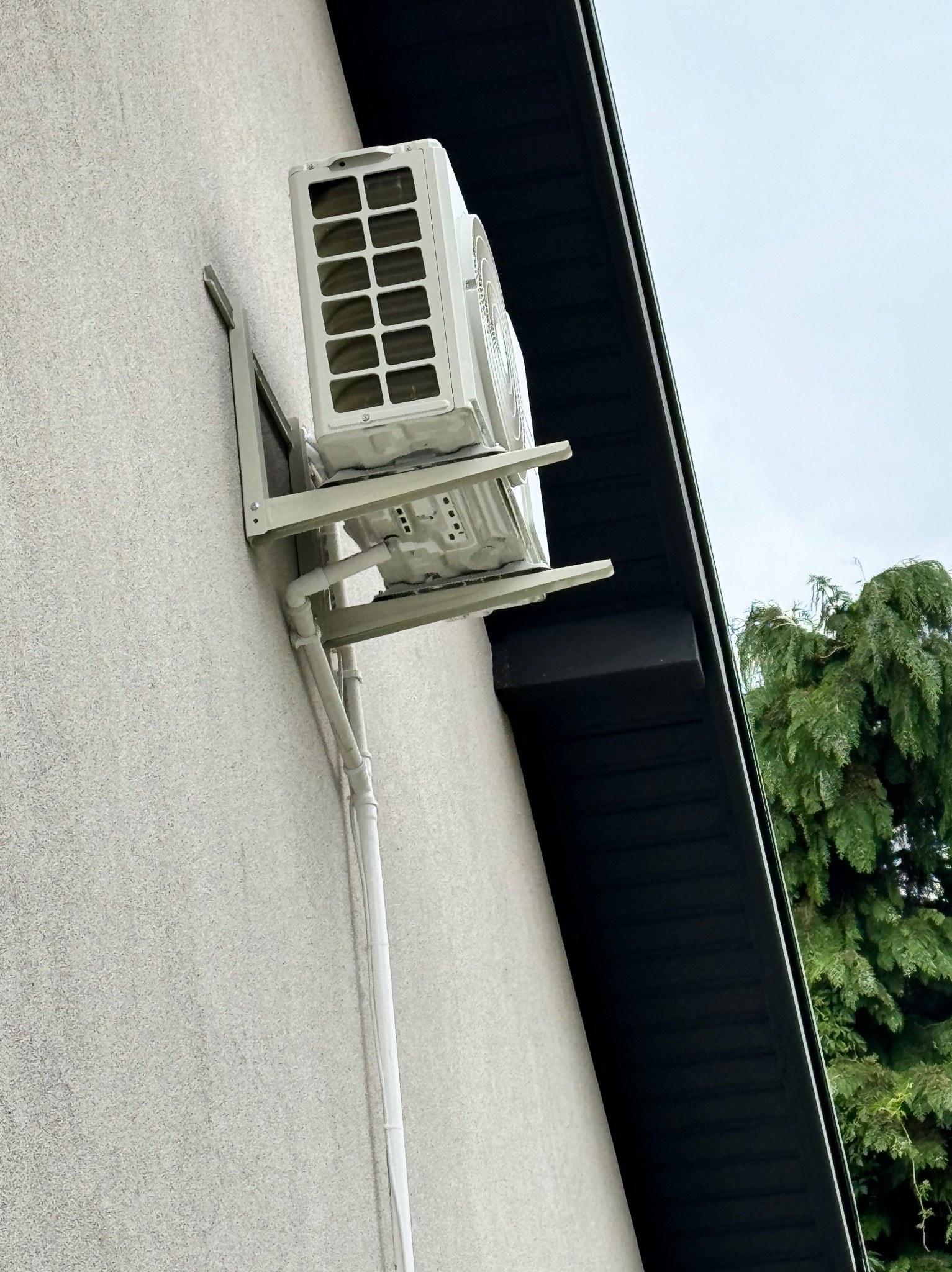 Zewnętrzna jednostka klimatyzacji zamontowana na ścianie domu z widocznymi rurami odprowadzającymi skropliny i fragmentem dachu oraz zielenią w tle.