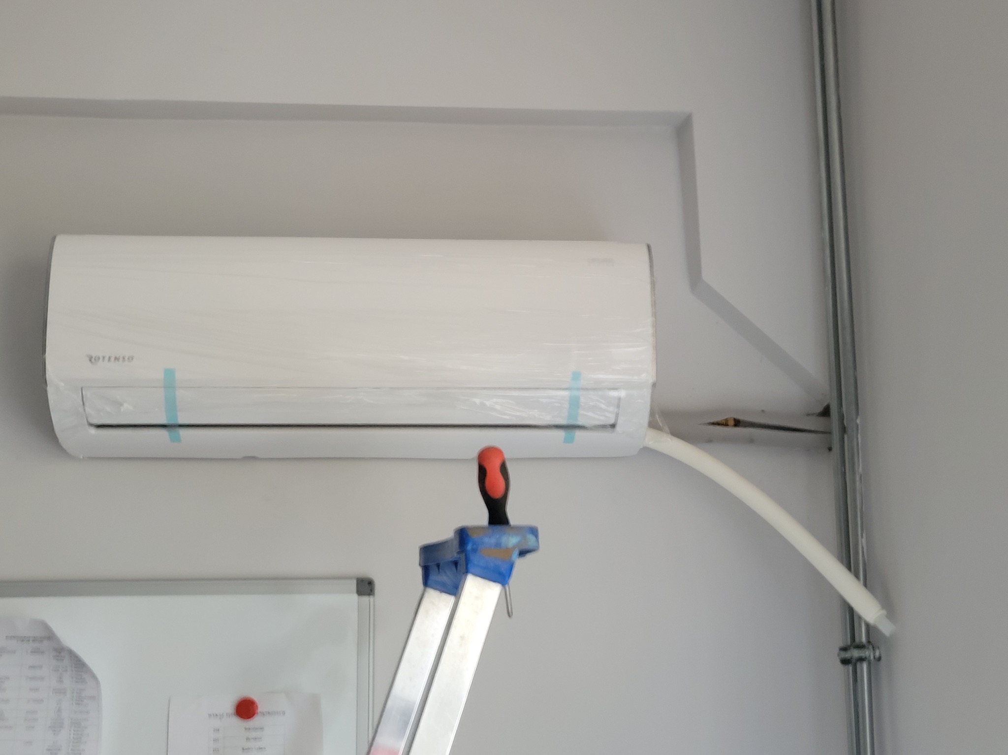 Biały klimatyzator ROTENSO, jeszcze w folii ochronnej, zamontowany na szarej ścianie, widoczna drabina z czerwono-czarnym śrubokrętem na górze oraz rurka odprowadzająca skropliny.