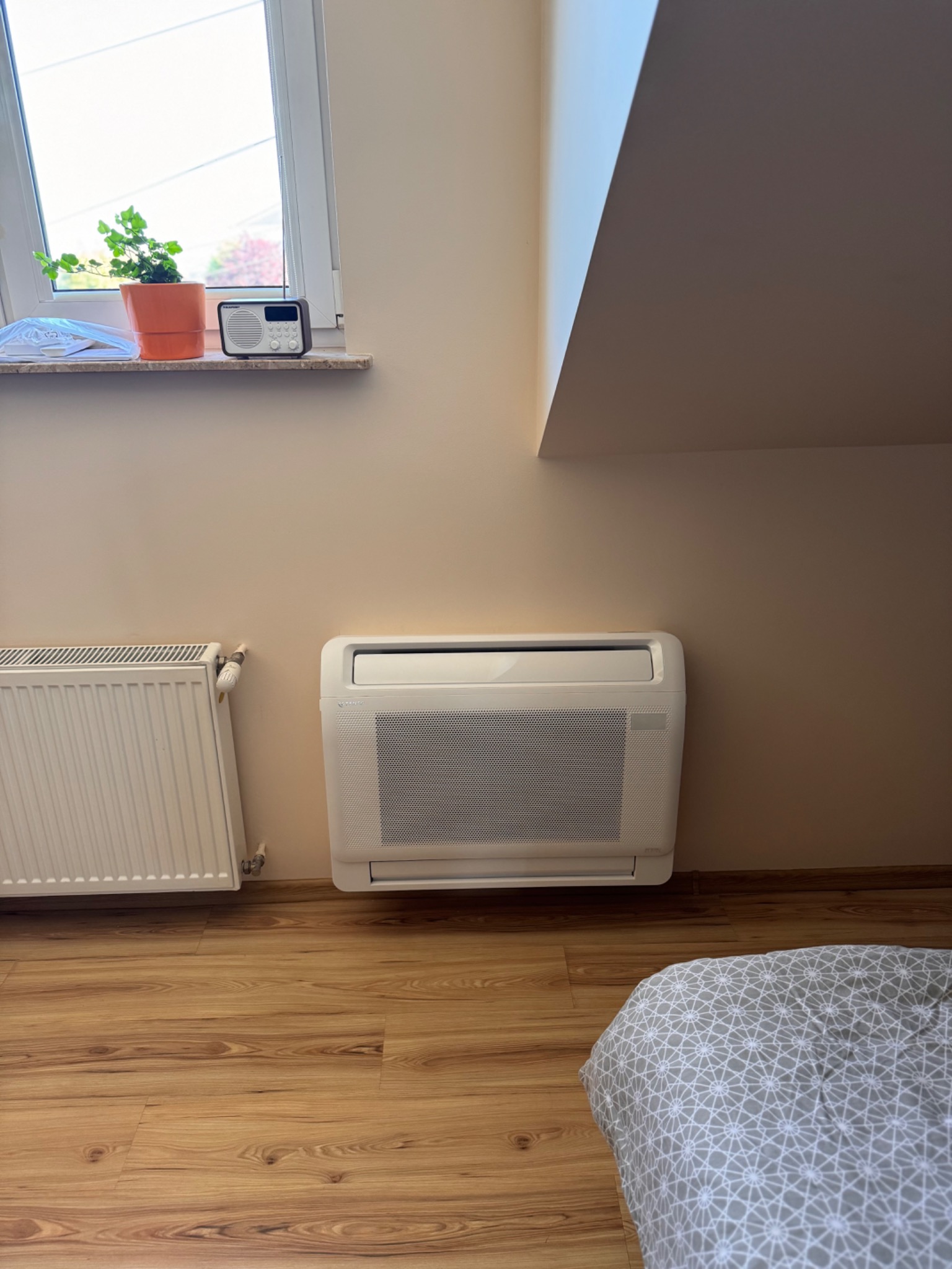 Klimatyzator podłogowy w jasnym pokoju na poddaszu, obok grzejnika i łóżka, z rośliną i radiem na parapecie okna.