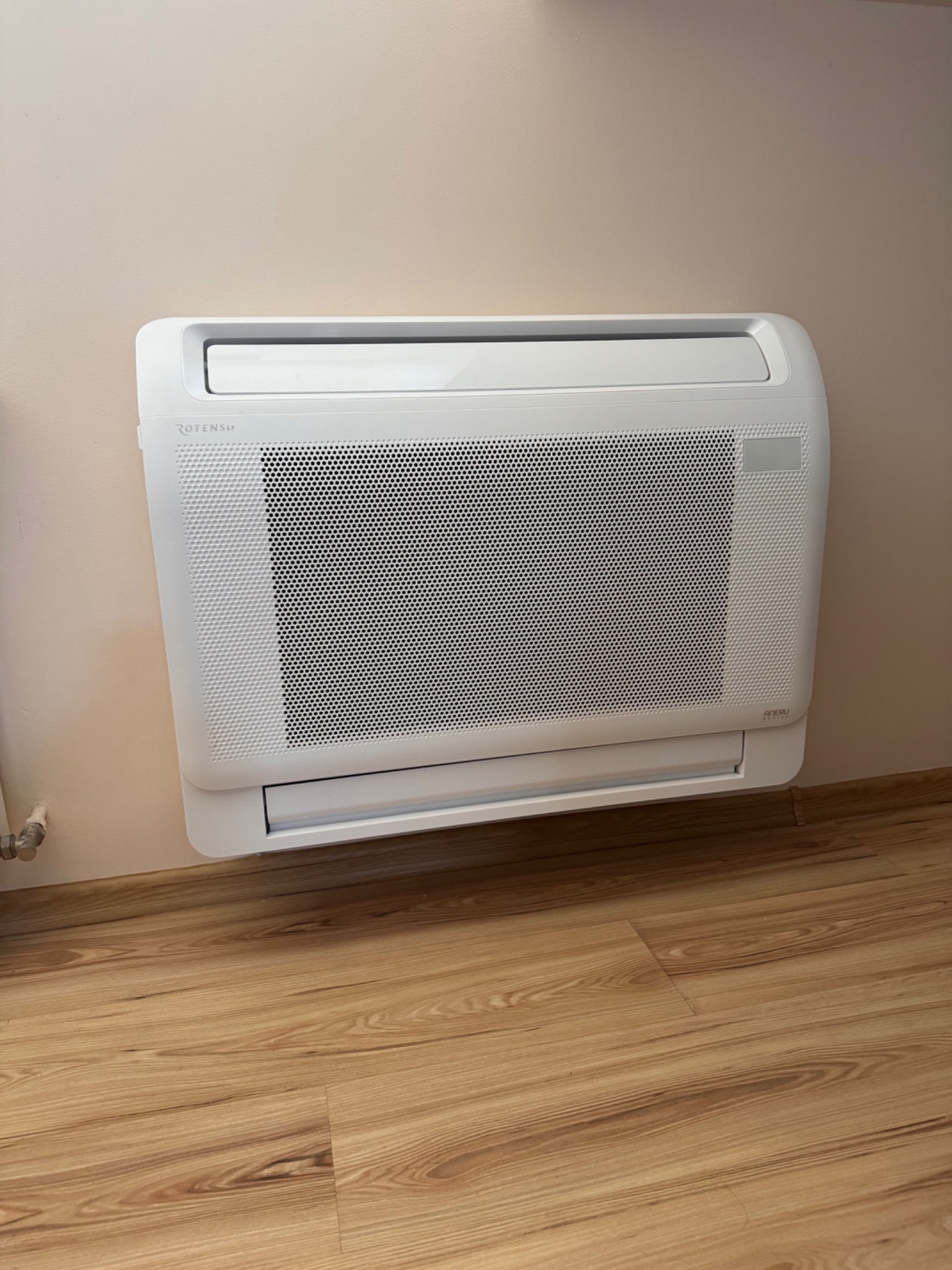 Biały klimakonwektor ROTENSO zamontowany na ścianie w pomieszczeniu z drewnianą podłogą.