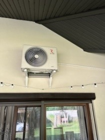 Biała jednostka klimatyzacji zamontowana na ścianie zewnętrznej budynku, widoczna z dołu, z dekoracyjnymi lampkami sznurkowymi poniżej i fragmentem rolety okiennej na pierwszym planie.