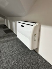 Biały klimatyzator ścienny zamontowany w pomieszczeniu ze skośnym sufitem i szarą wykładziną dywanową.