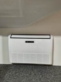 Biała jednostka wewnętrzna klimatyzacji przypodłogowej zamontowana pod skosem dachu na szarej wykładzinie, widoczne kratki wylotowe i panel sterowania.