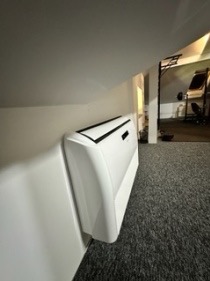 Biała jednostka wewnętrzna klimatyzacji ściennej zamontowana pod skosem dachu na szarej wykładzinie dywanowej.