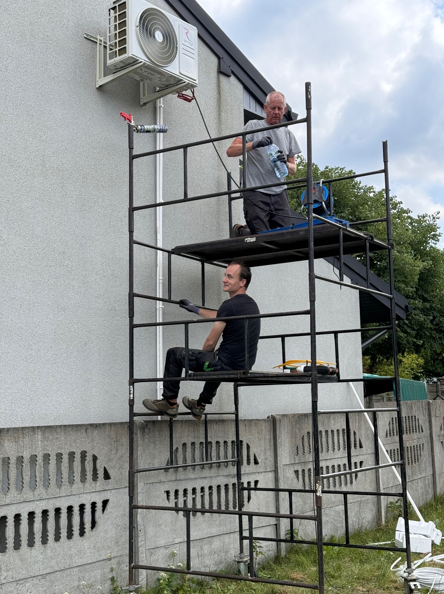 Dwóch monterów na rusztowaniu podczas instalacji klimatyzatora na ścianie domu; jeden z nich pije wodę, drugi siedzi i patrzy w dal; widoczny klimatyzator na elewacji budynku.