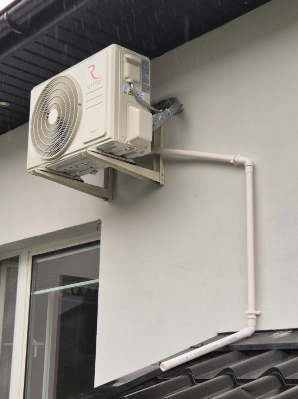 Biała jednostka zewnętrzna klimatyzacji marki Rotenso zamontowana na jasnej ścianie budynku, widoczne białe rury odprowadzające skropliny na czarny dach, instalacja przy oknie, pochmurna pogoda.