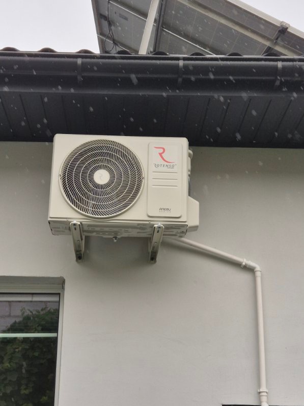 Zewnętrzna jednostka klimatyzacji Rotenso zamontowana na ścianie domu, widoczne rury odprowadzające skropliny i panel słoneczny na dachu w tle.