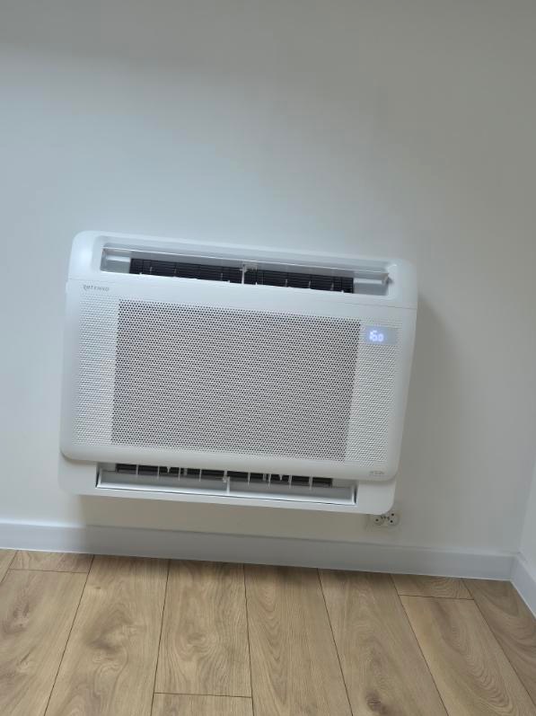 Biały klimakonwektor ścienny z widocznym wyświetlaczem temperatury, zamontowany w pomieszczeniu biurowym z panelami podłogowymi.