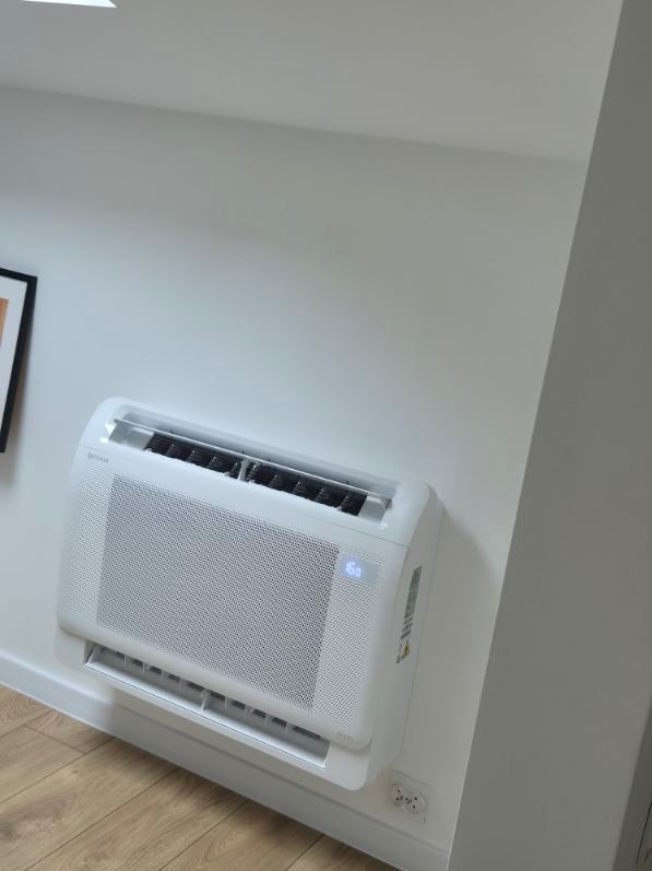 Biały klimatyzator ścienny z wyświetlaczem cyfrowym, zamontowany w nowoczesnym wnętrzu z drewnianą podłogą, widoczne gniazdko elektryczne w ścianie.