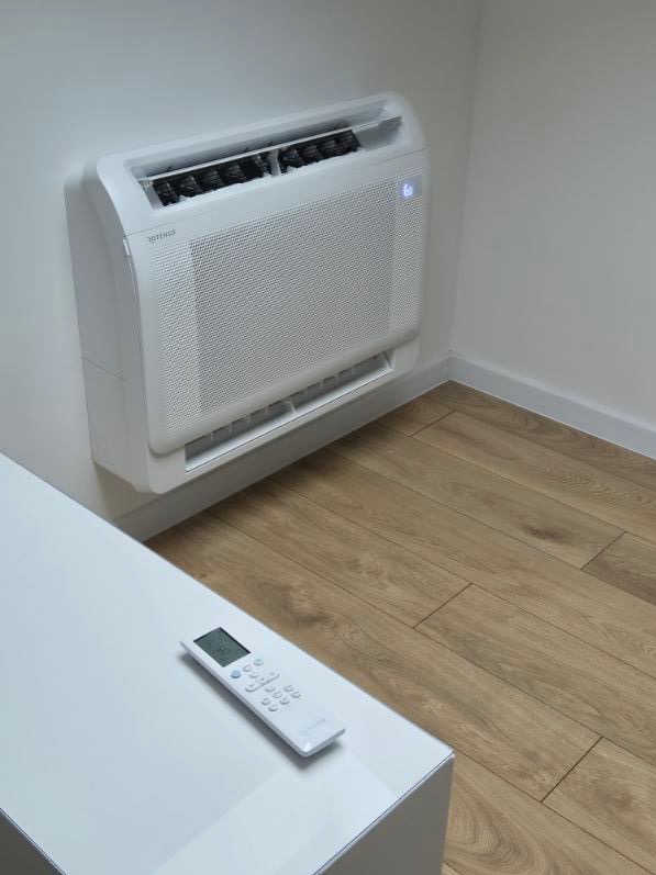 Biała jednostka klimatyzacji podłogowej zamontowana na ścianie w rogu pomieszczenia, obok leży pilot na białym blacie, widoczna drewniana podłoga.