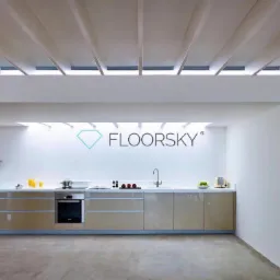 Nowoczesna kuchnia z beżowymi szafkami na tle białej ściany z logo firmy Floorsky, widoczne oświetlenie sufitowe i betonowa posadzka.