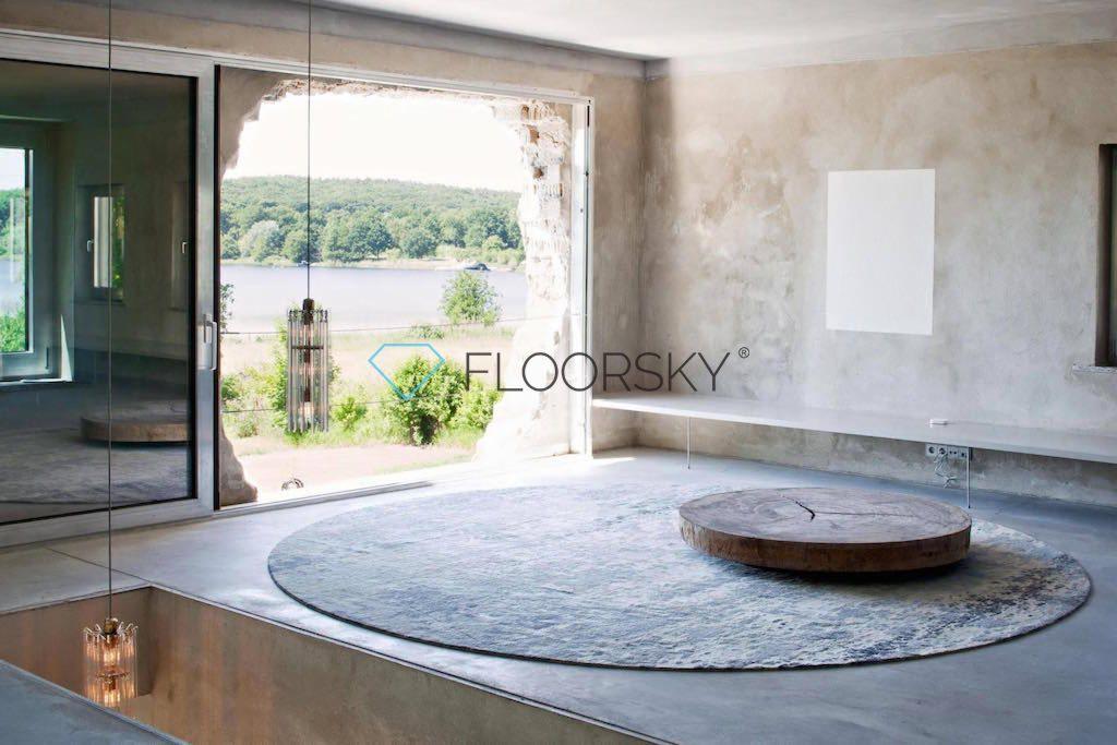 Wnętrze z posadzką w odcieniach szarości, okrągłym dywanem i stolikiem z przekroju pnia drzewa, widok na jezioro przez nieregularny otwór okienny, minimalistyczny design.