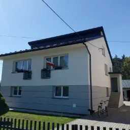 Dwukondygnacyjny dom jednorodzinny z jasną elewacją podzieloną poziomym pasem szarości, z polską flagą w oknie, rowerami opartymi o ścianę i psem na podwórku.