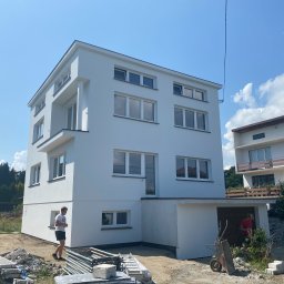 Dawid Smolik, Usługi Budowlane - Nowoczesny, trzypiętrowy dom z białą elewacją w słoneczny dzień. Widoczne rusztowania i pracownik, co sugeruje prace wykończeniowe. Minimalistyczny design budynku.