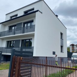 Dawid Smolik, Usługi Budowlane - Nowoczesny, dwupiętrowy dom z białą elewacją i szarymi balkonami, widziany zza brązowego ogrodzenia. Na tle pochmurne niebo i częściowo widoczny samochód dostawczy.