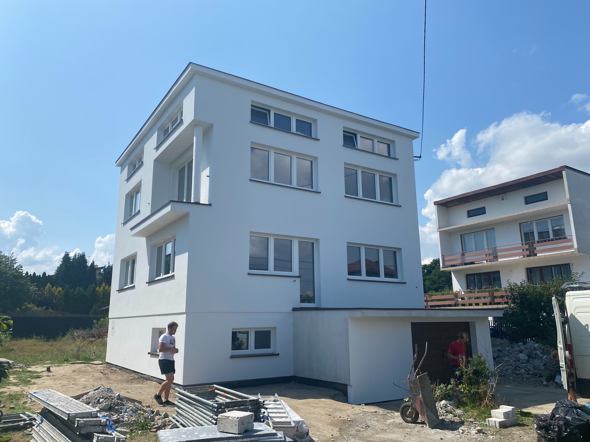 Nowoczesny, trzypiętrowy dom z białą elewacją w słoneczny dzień. Widoczne rusztowania i pracownik, co sugeruje prace wykończeniowe. Minimalistyczny design budynku.