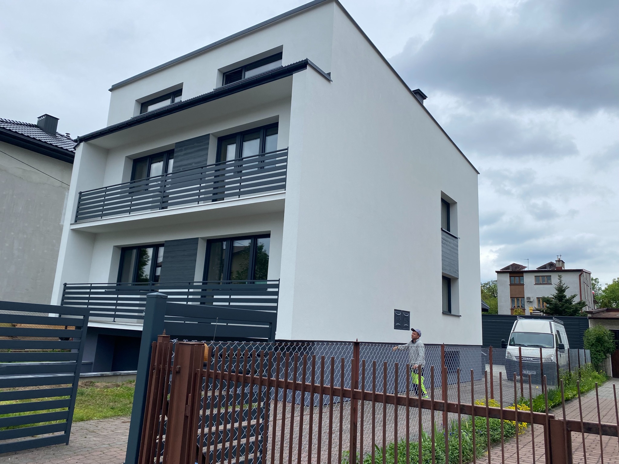 Nowoczesny, dwupiętrowy dom z białą elewacją i szarymi balkonami, widziany zza brązowego ogrodzenia. Na tle pochmurne niebo i częściowo widoczny samochód dostawczy.
