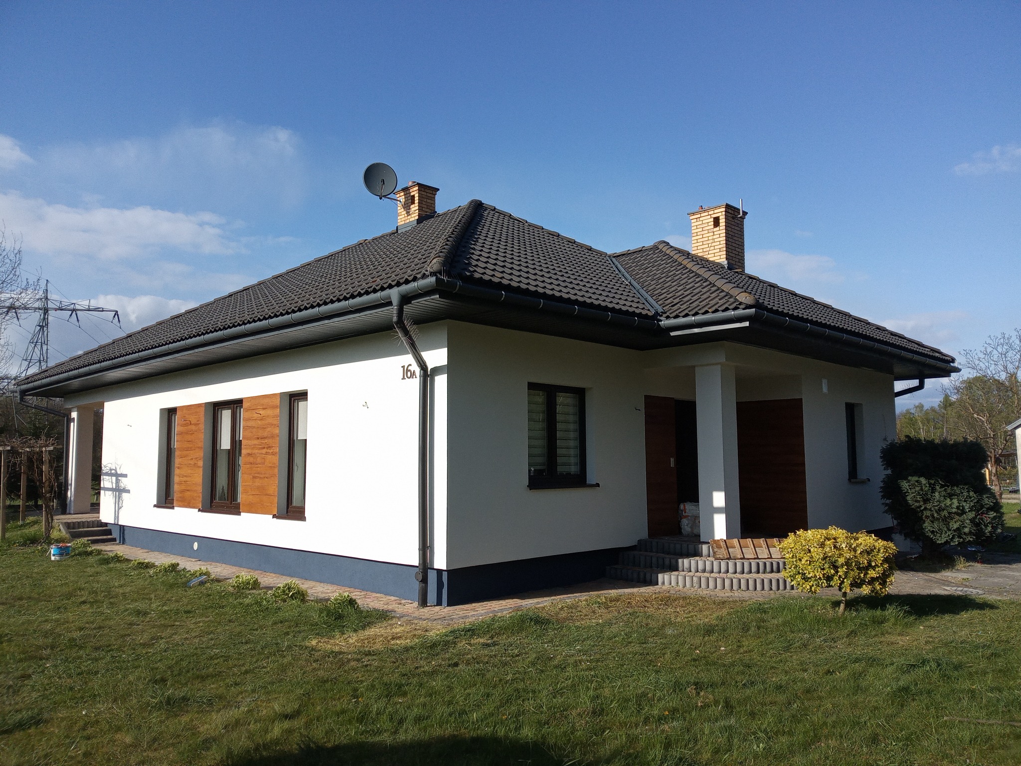 Parterowy dom z białą elewacją i drewnianymi okiennicami, ciemny dach z kominami, satelita. Widok z ogrodu w słoneczny dzień, błękitne niebo.