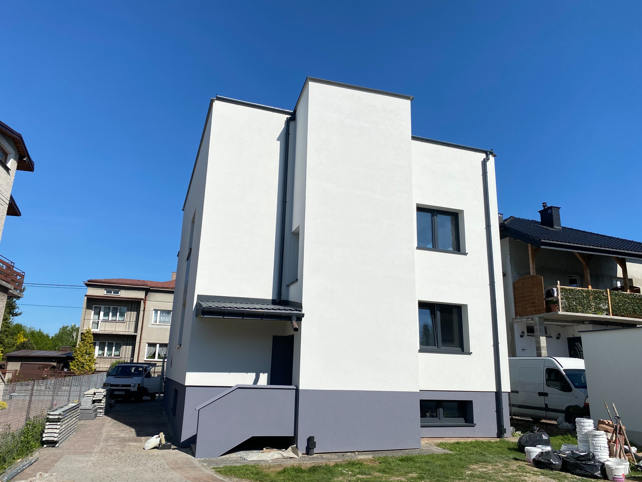 Nowoczesny, dwukondygnacyjny dom z białą elewacją i szarym cokołem, widoczny z perspektywy trawnika. W tle inne budynki i błękitne niebo.