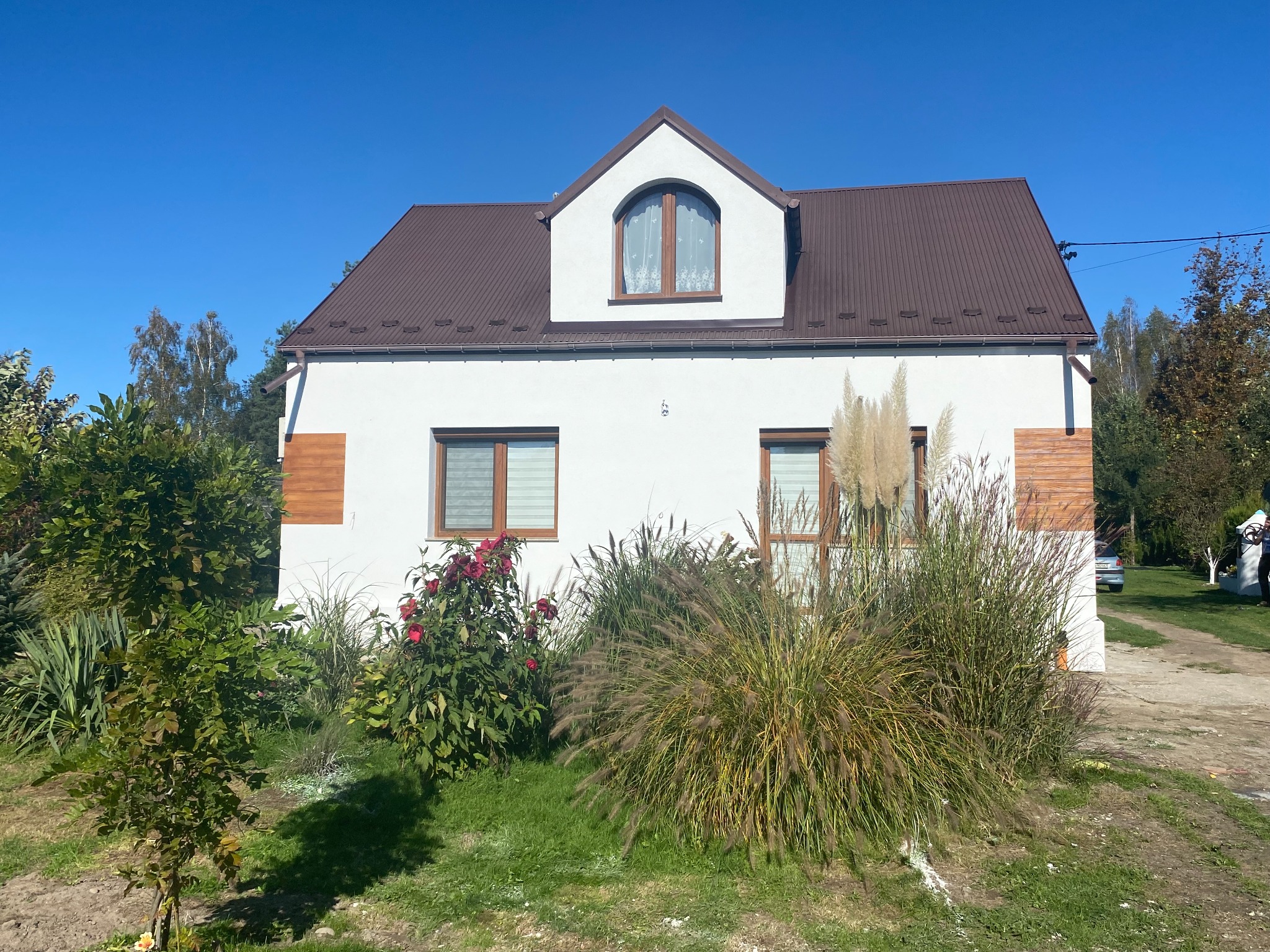 Biały dom jednorodzinny z brązowym dachem i drewnianymi akcentami elewacji, otoczony zielenią w słoneczny dzień. Widoczne okna z roletami i roślinność ozdobna.