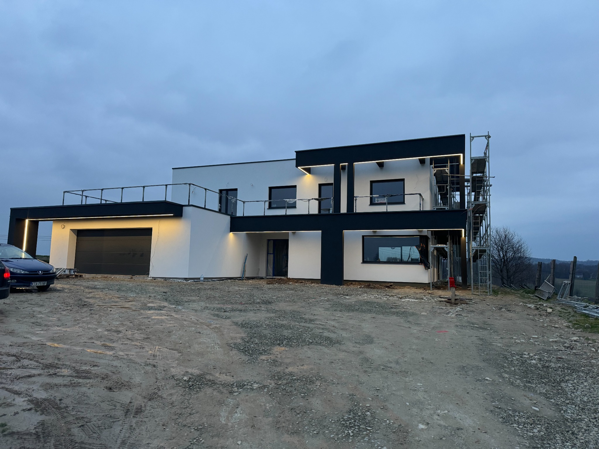 Nowoczesny dom z białą elewacją i czarnymi akcentami, oświetleniem zewnętrznym i rusztowaniami. Widok na posesję w trakcie prac wykończeniowych.