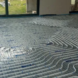 Białe rury grzewcze ułożone w pętle na folii izolacyjnej z siatką, przygotowane do wylewki pod ogrzewanie podłogowe, widok na okno i trawnik w tle.