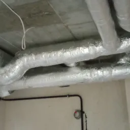 Srebrne, zaizolowane rury wentylacyjne pod betonowym sufitem, widoczne czarne rury instalacji elektrycznej wzdłuż ściany.