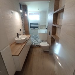 KRISBUD KRZYSZTOF DOROTA STASZEK - Nowoczesna łazienka z umywalką nablatową na drewnianym blacie, białą wanną wolnostojącą, toaletą i grzejnikiem drabinkowym, wykończona płytkami w odcieniach beżu i brązu z elementami dekoracyjnymi.